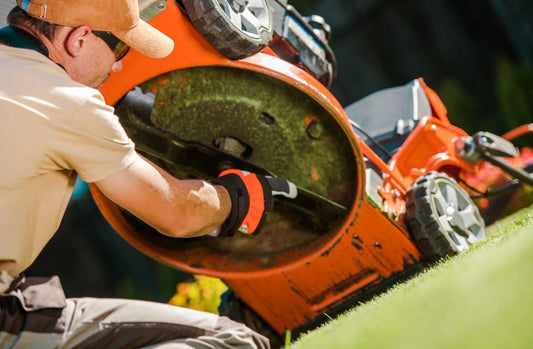 A man using an orange lawn mower to cut grass, removing the mower's sharp blade for maintanence.